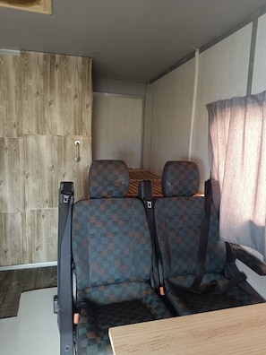 Ein kompaktes Wohnmobil-Interieur mit zwei Sitzgruppen, einer Wand aus Holzpaneelen und einem Fenster mit transparenten Vorhängen.