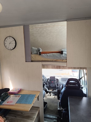 Ein kompaktes Wohnmobil-Interieur mit einer Uhr, einem Bett mit gemusterten Betttextilien, einem Tisch mit einem Notizbuch und einer Schachtel sowie einem Lenkrad.