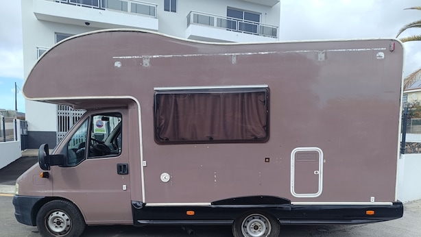 Ruskea asuntoauto on pysäköitynä rakennuksen eteen, ja rakennuksessa on parveke.