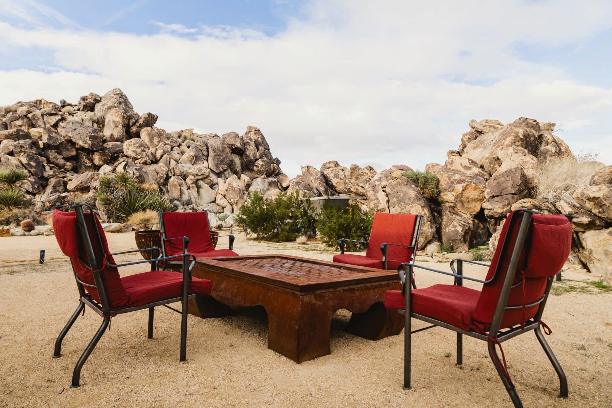 An outdoor seating area with a metal table and chairs, set against a backdrop of large rocks and sparse vegetation.