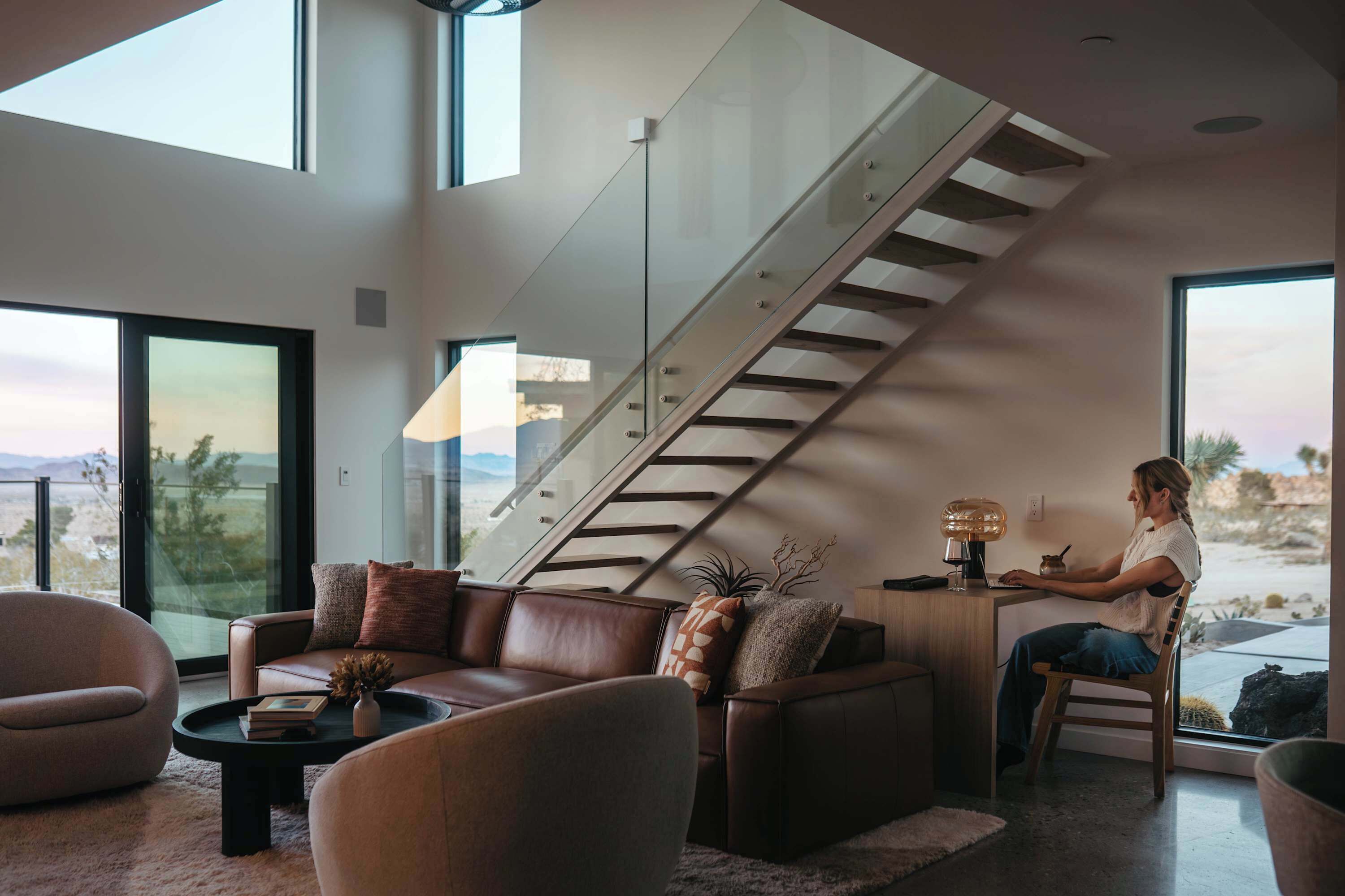 A modern living room with a staircase, large windows, and a person working at a desk.