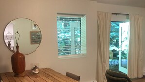 Un coin repas avec une table en bois, des chaises, un vase et un miroir. Il y a une fenêtre avec des stores et une vue sur la verdure à l’extérieur.
