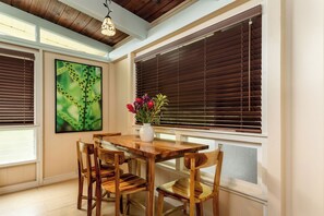 A dining area with a wooden table and chairs, a vase with flowers, and a large window with blinds.
