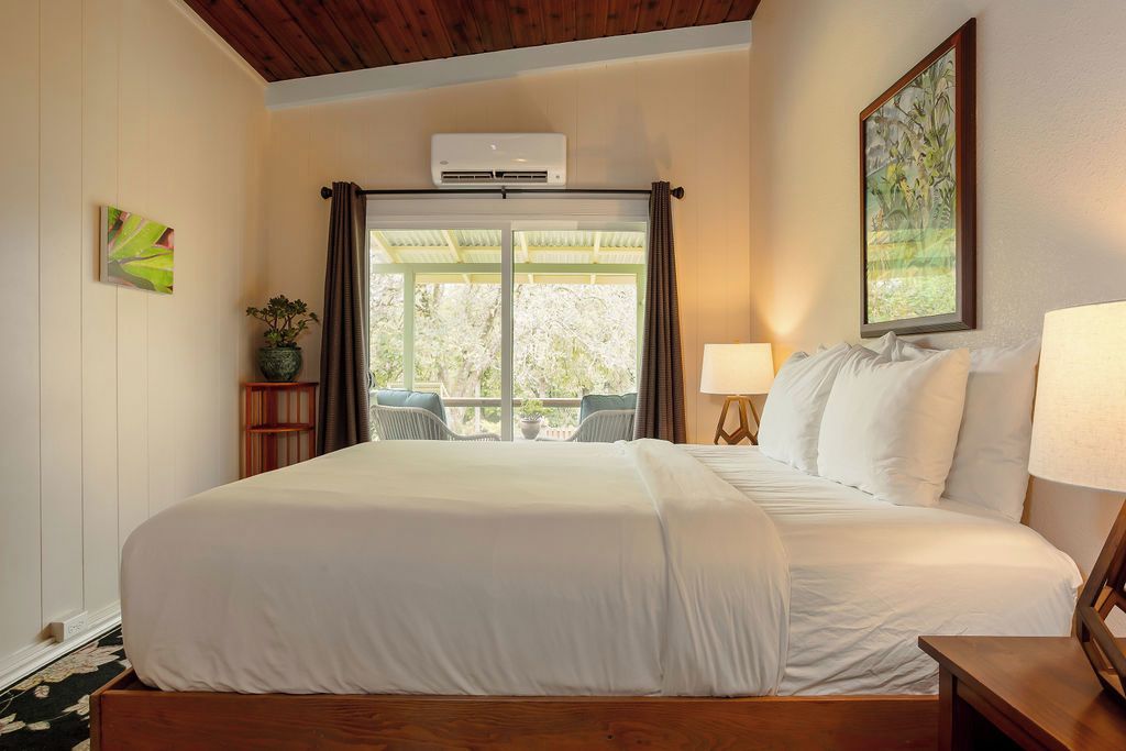 A hotel room with a large bed, a window with curtains, a wall-mounted air conditioner, a framed picture, and a small table with a lamp.