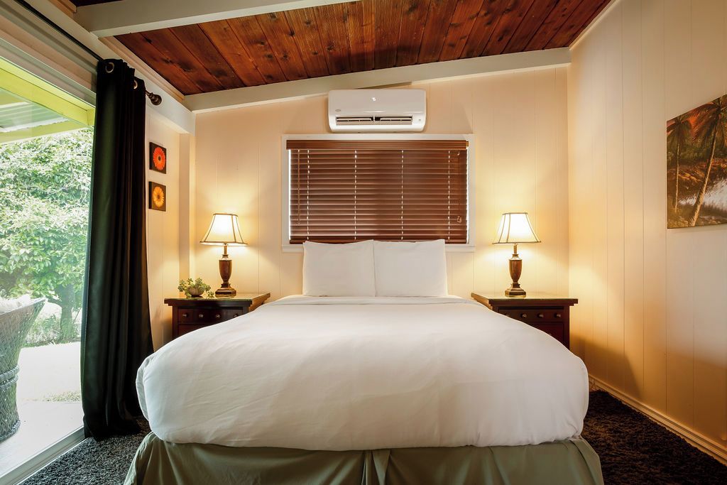 A hotel room with a bed, two bedside lamps, a window with blinds, an air conditioning unit, and a painting on the wall.