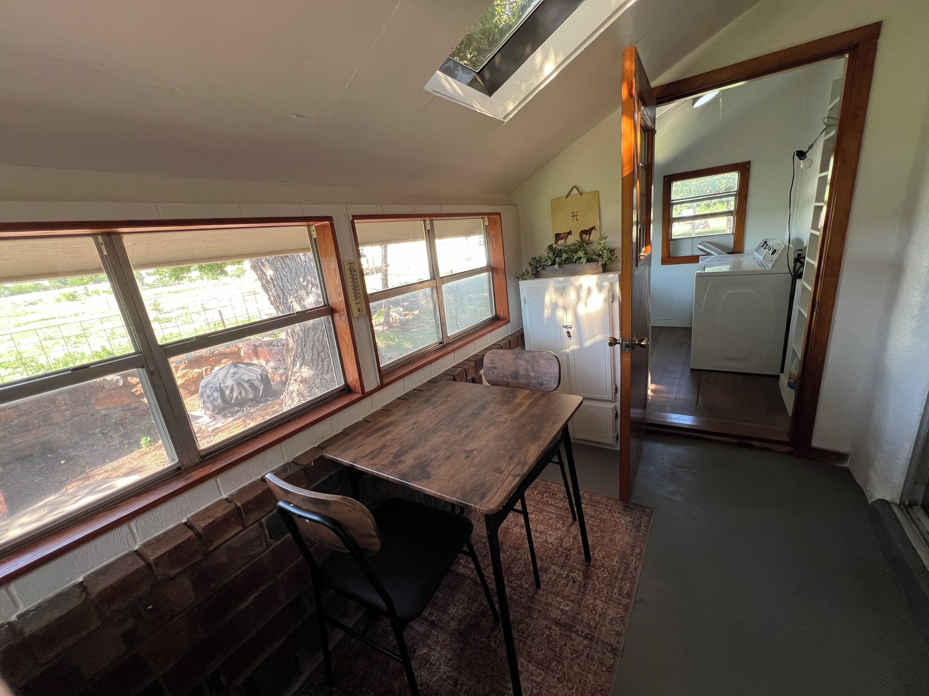 Une pièce comprenant une table et des chaises, une fenêtre donnant sur un espace extérieur, un meuble blanc orné d’une plante, et une porte menant à la cuisine.