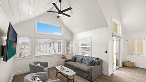 En rummelig stue med en grå sofa, et sofabord og en loftventilator. Store vinduer med skodder, et vægmonteret tv og en dør, der fører til et andet rum.