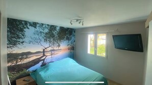 Ett sovrum med en stor väggmålning som föreställer ett träd och ett landskap, en säng med ett turkosfärgat överkast, ett sängbord, ett fönster som släpper in dagsljus och en väggmonterad TV.
