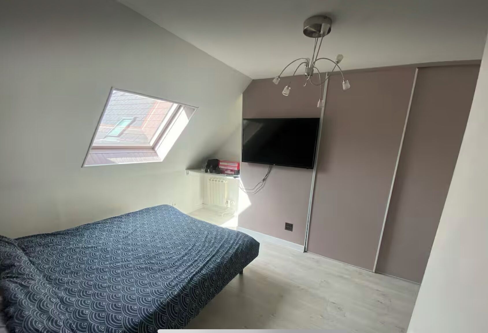 A bedroom with a bed, a TV mounted on the wall, a window, and a ceiling light fixture.