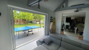 Ett vardagsrum med en grå soffa, ett bordtennisbord och utsikt över en trädgård med ett trädäck och grönska.