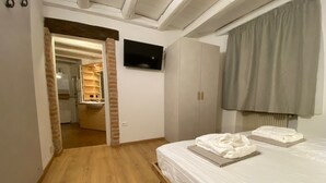 A bedroom with a wooden floor, a bed with white linens, a TV on the wall, and a bathroom visible through an open door.