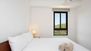 Habitación de hotel con cama, mesita de noche con lámpara y ventana con vista a follaje.