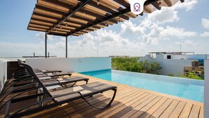 Zona de piscina en la azotea con terraza de madera y sillones de descanso, con vistas a un paisaje urbano y zonas verdes.