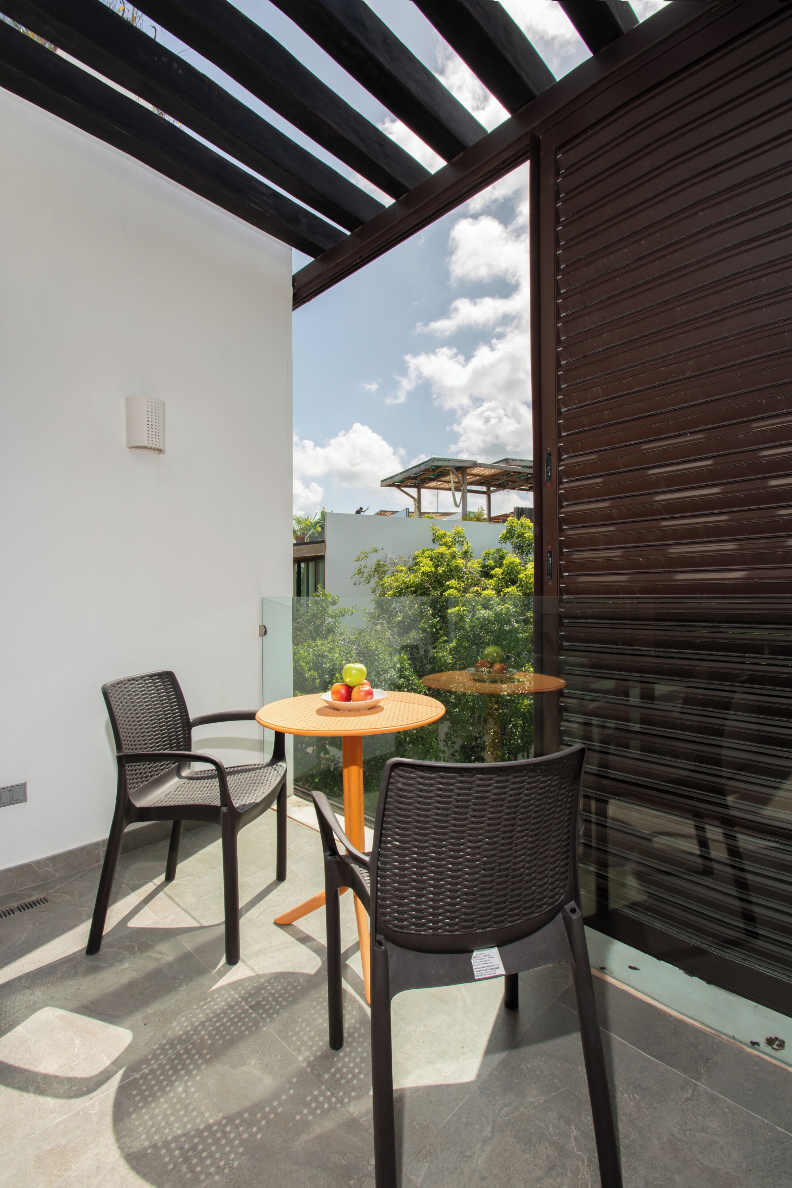 A balcony with a round table and two chairs, a view of greenery, and a closed shutter.