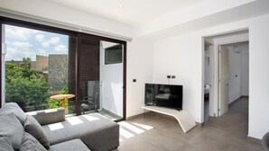 Una sala de estar moderna con un sofá gris, un televisor de pantalla plana sobre un mueble bajo y un ventanal corredizo que da a un balcón con mesa y sillas.