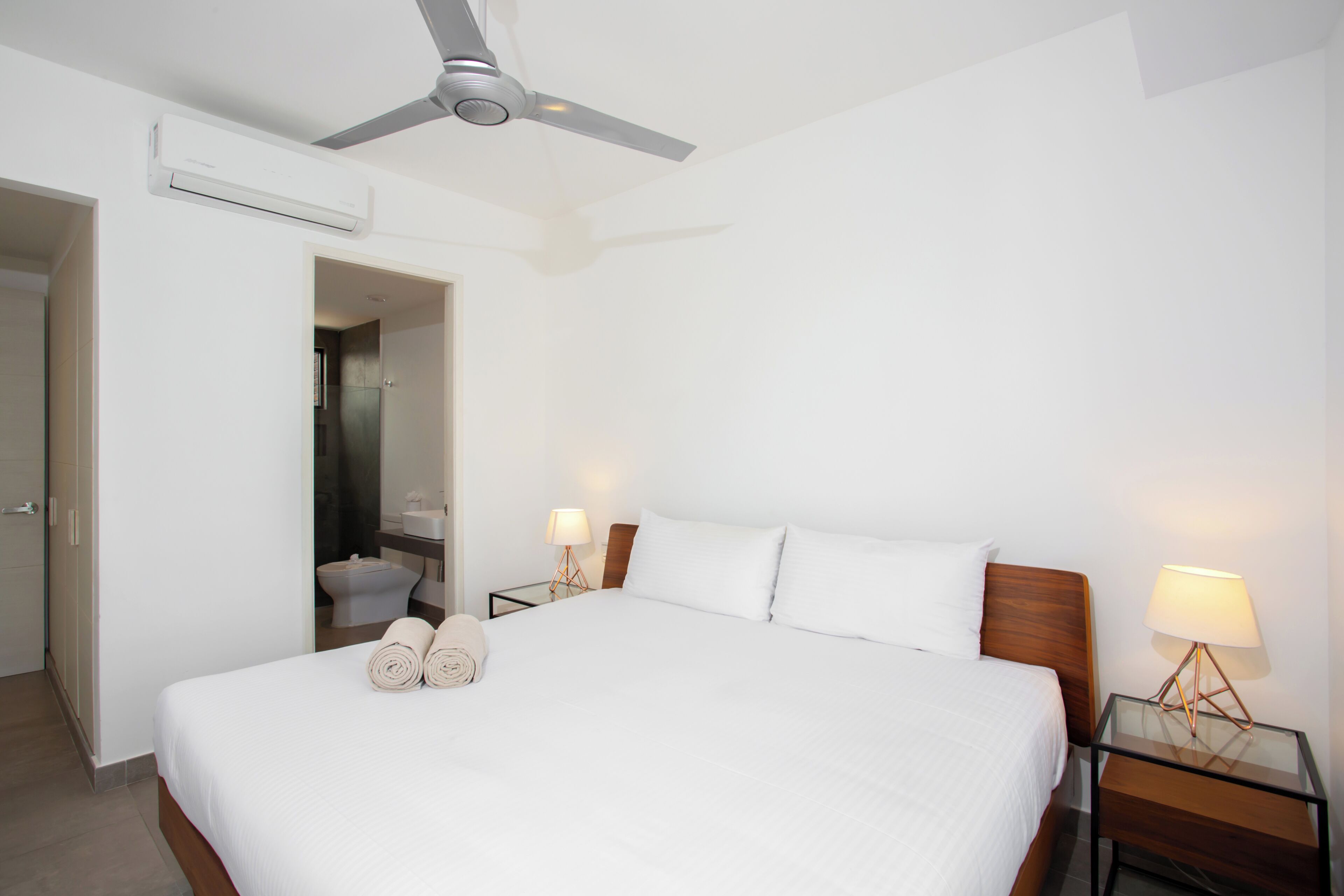 Una habitación de hotel con una cama, dos mesitas de noche con lámparas, un ventilador de techo, un equipo de aire acondicionado y un baño visible a través de una puerta abierta.