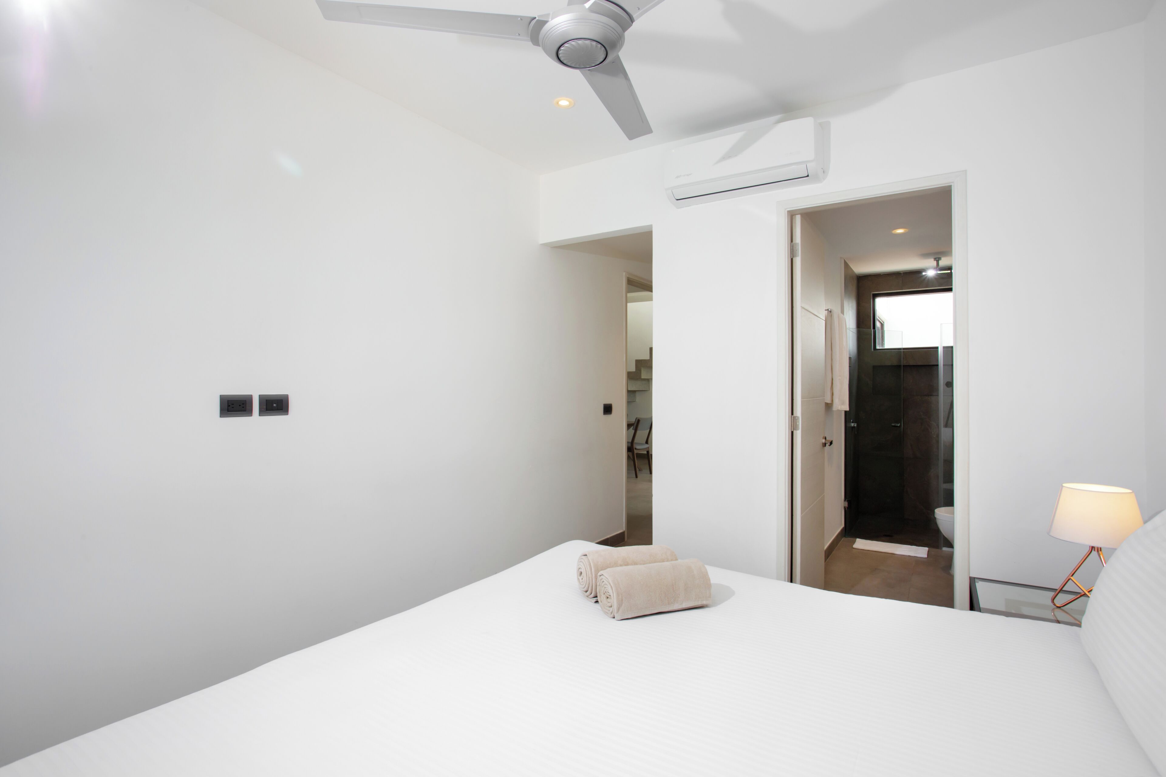 A modern hotel room with a bed, a ceiling fan, a bedside lamp, and a view into the bathroom.