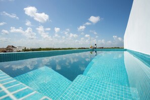 Une piscine d’un bleu éclatant, bordée de carreaux, d’où l’on peut admirer le ciel et les bâtiments lointains.