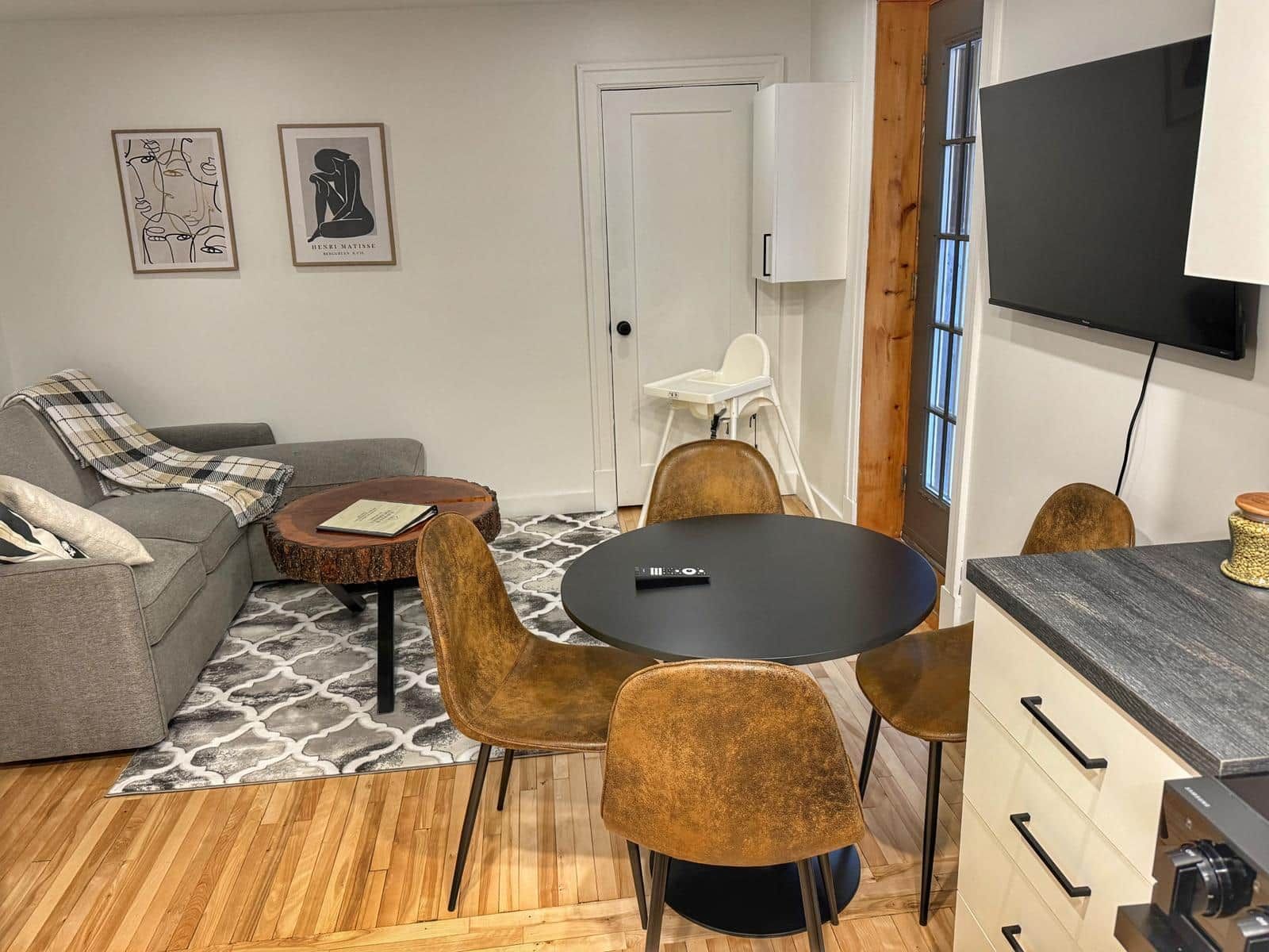 A modern living room with a grey sofa, a round table, and two chairs. There is a white door, a white cabinet, and a flat-screen TV mounted on the wall.