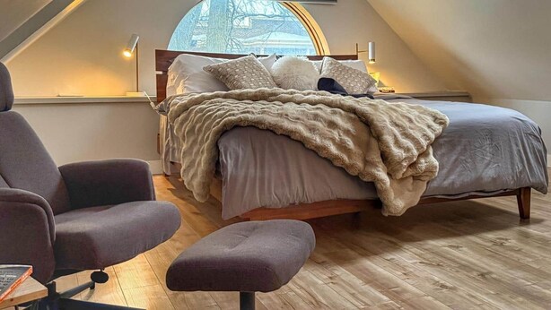Um quarto aconchegante com teto inclinado, uma janela grande, uma cama com cobertor macio e duas cadeiras.