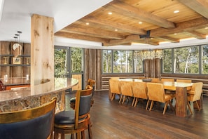 Un vaste espace dédié à la salle à manger, avec une grande table et des chaises en bois, un coin bar doté d’un comptoir en marbre et de grandes fenêtres offrant une vue sur l’extérieur.