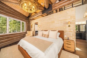 Une chambre avec un grand lit, des murs en pierre, un plafond en bois et une vue sur les arbres par la fenêtre.