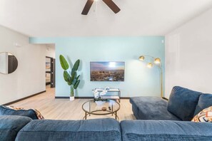 Una sala de estar con sofá, mesa de centro, planta, cuadro y lámpara de pie.