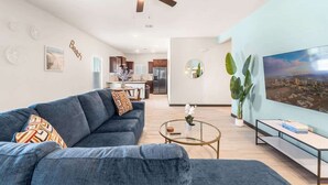 Un soggiorno con divano, tavolino e televisore. Si nota un ventilatore a soffitto, una cucina sullo sfondo e un'ampia finestra con vista sulla città.