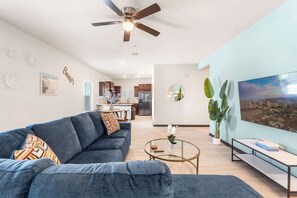 Una sala de estar con sofá, mesa de centro y televisor. Hay un ventilador de techo, una cocina al fondo y un ventanal con vista a la ciudad.