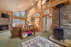 A cozy cabin interior with a stone fireplace, a wooden sofa, a table with a basket, and a black wood-burning stove. - Serene Mountain Retreat with Private Beach Access (McCall)