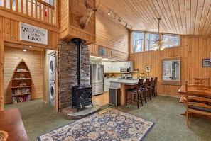 A cabin interior with a stone fireplace, kitchen, dining area, and a 'Welcome to the Lake' sign. - Serene Mountain Retreat with Private Beach Access (McCall)