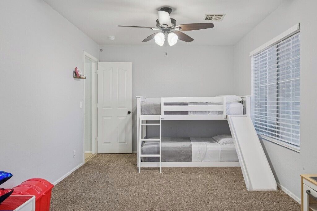 Une petite pièce bien éclairée, avec un lit superposé, un ventilateur de plafond et une fenêtre équipée de stores.