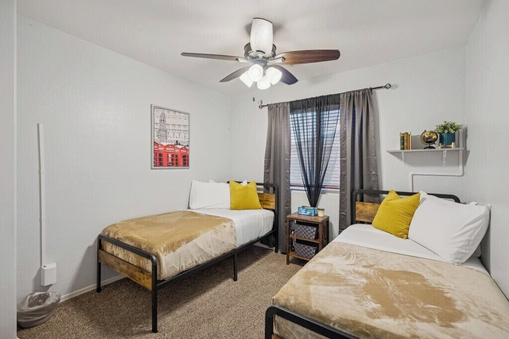 A room with two beds, a ceiling fan, a window with curtains, a small table with a globe, and a shelf with books and a plant.
