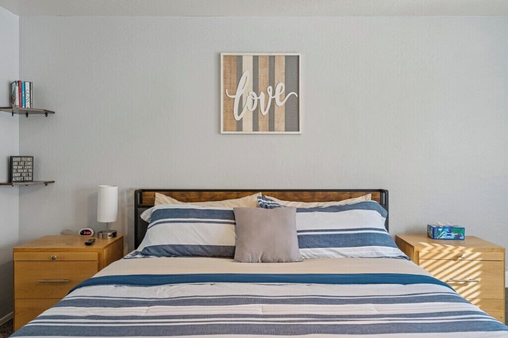 Ein ordentlich eingerichtetes Schlafzimmer mit einem Bett, Kissen, einem Nachttisch, einer Wandablage und einem gerahmten Kunstwerk.