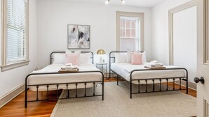 A bedroom with two beds, white walls, a ceiling light fixture, and a window with blinds.