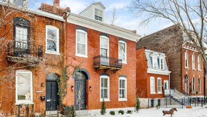 A red brick townhouse with a black front door, white shutters, and a small balcony.