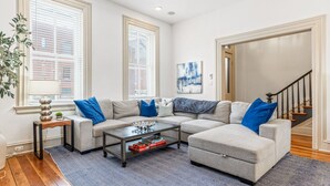 A living room with a sectional sofa, a coffee table, and a rug. There are two windows with blinds, a painting on the wall, and a staircase in the background.