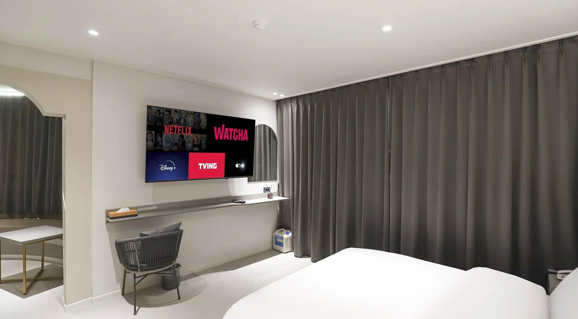 A modern hotel room with a large flat-screen TV displaying various streaming services, a comfortable bed, a small desk with a chair, and a large window with curtains.