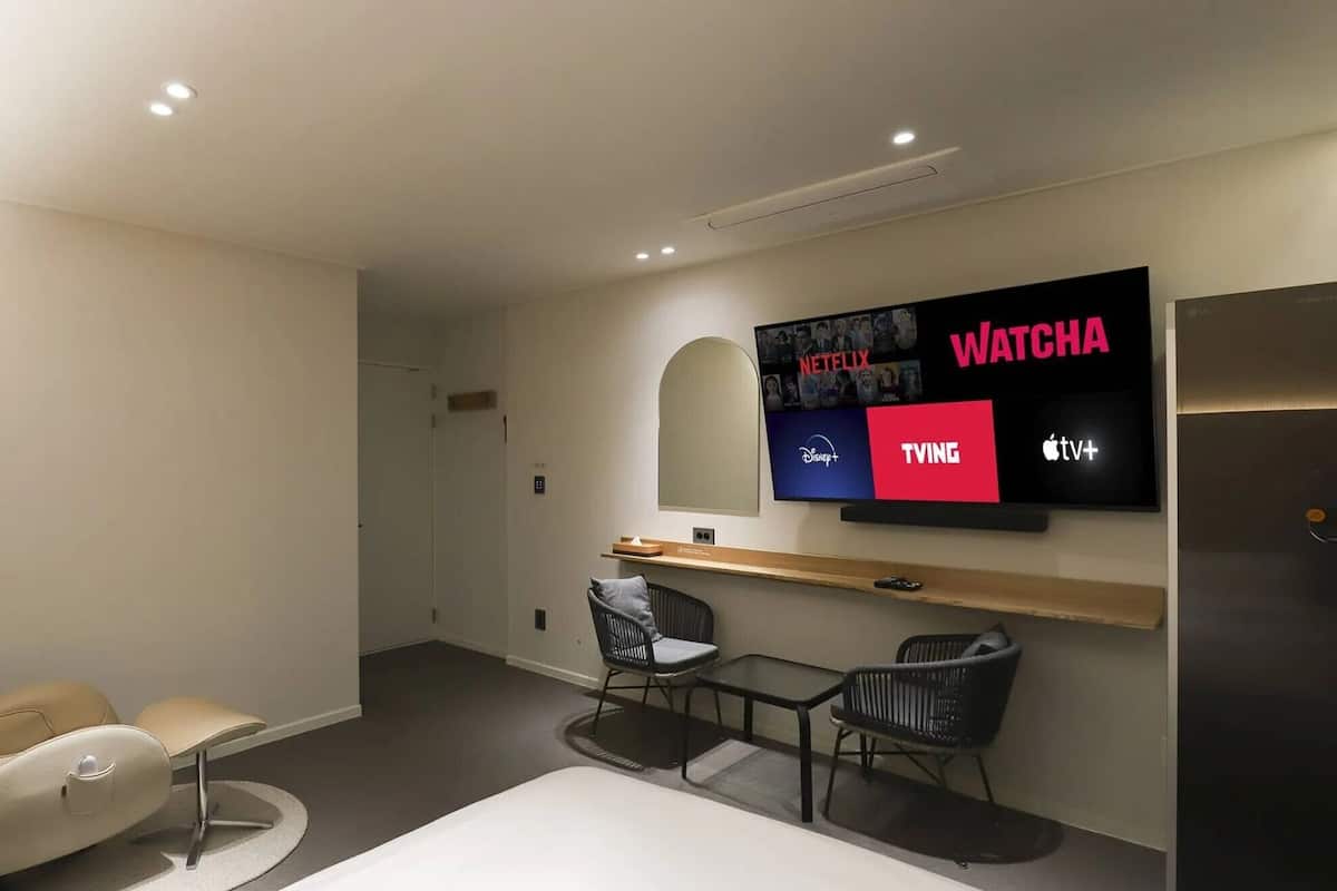 Une chambre d’hôtel moderne dotée d’un grand téléviseur à écran plat qui permet d’accéder à divers services de diffusion en continu, d’un fauteuil massant, d’une petite table ronde et d’un design minimaliste.