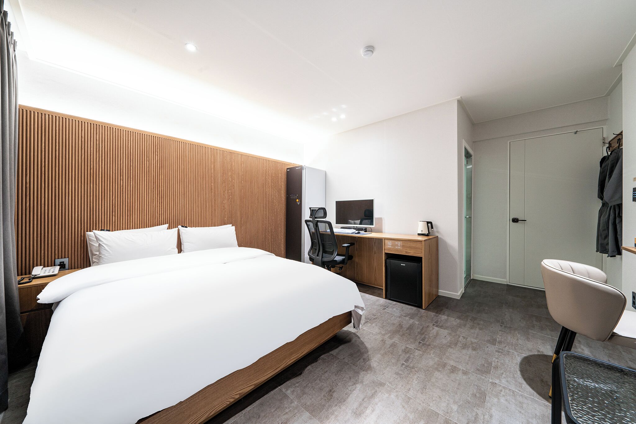 Una habitación de hotel moderna con una cama grande, cabecera de madera, un escritorio con computadora, una silla y un armario.