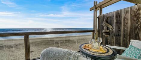 A balcony with a view of the beach, a table with snacks and drinks, and a wooden wall with a carved bird.