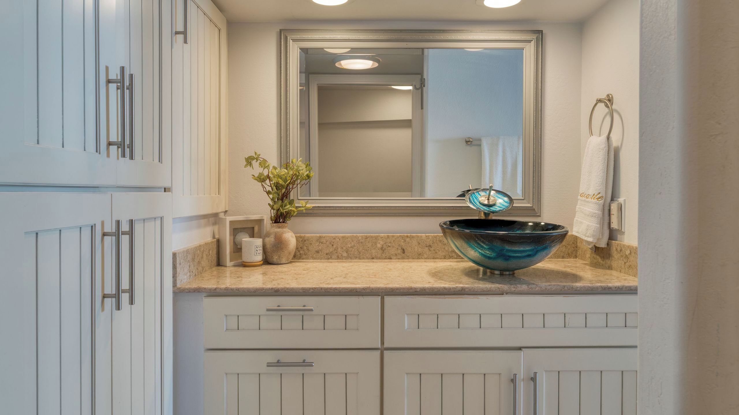 Une salle de bain avec un grand miroir, un lavabo agrémenté d’un récipient décoratif, une plante en pot et un porte-serviettes.