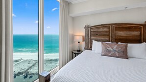 Hotelkamer met groot raam met uitzicht op een strand, houten hoofdeinde en een kussen met de tekst 'Seas the Day'.