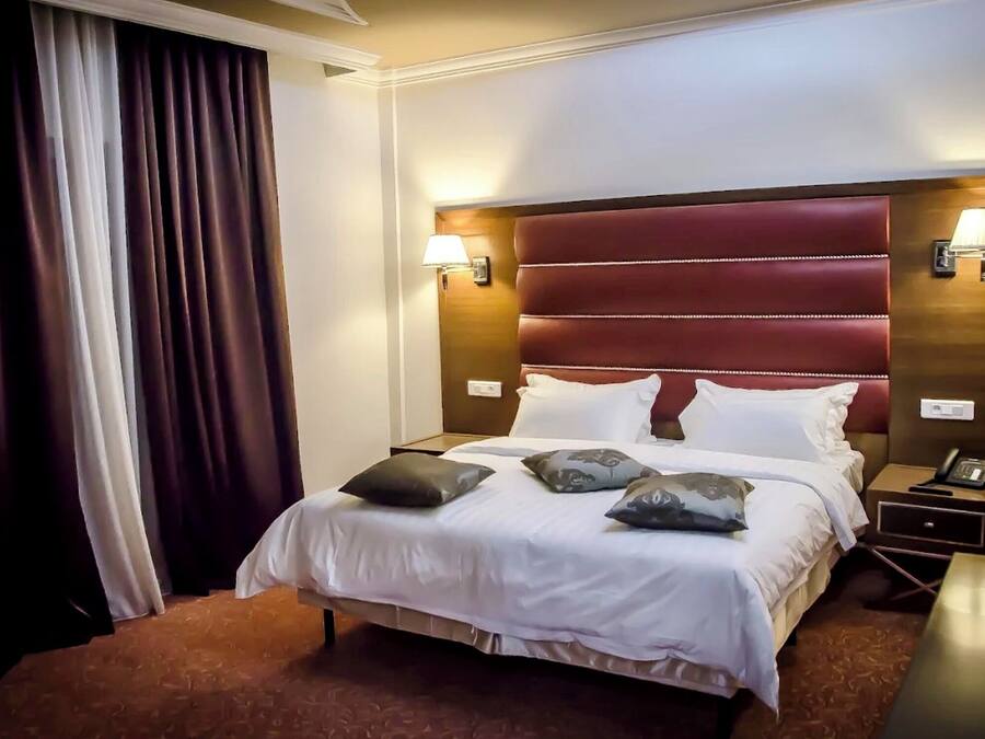 A hotel room with a large bed, two bedside lamps, a nightstand, and a window with curtains.