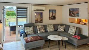 Un salon moderne avec un canapé d’angle gris, une petite table blanche et un banc recouvert d’une couverture. Trois tableaux encadrés ornent le mur, et un appareil de climatisation est installé au-dessus du canapé.