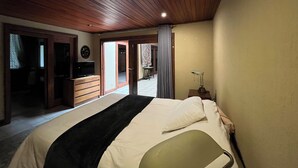Quarto com teto de madeira, cama com manta preta, abajur e televisão sobre uma cômoda de madeira.
