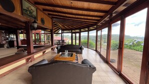 Uma sala de estar ampla com teto de madeira, um sofá grande, uma mesa de centro e vista para uma paisagem exuberante através de janelas amplas.