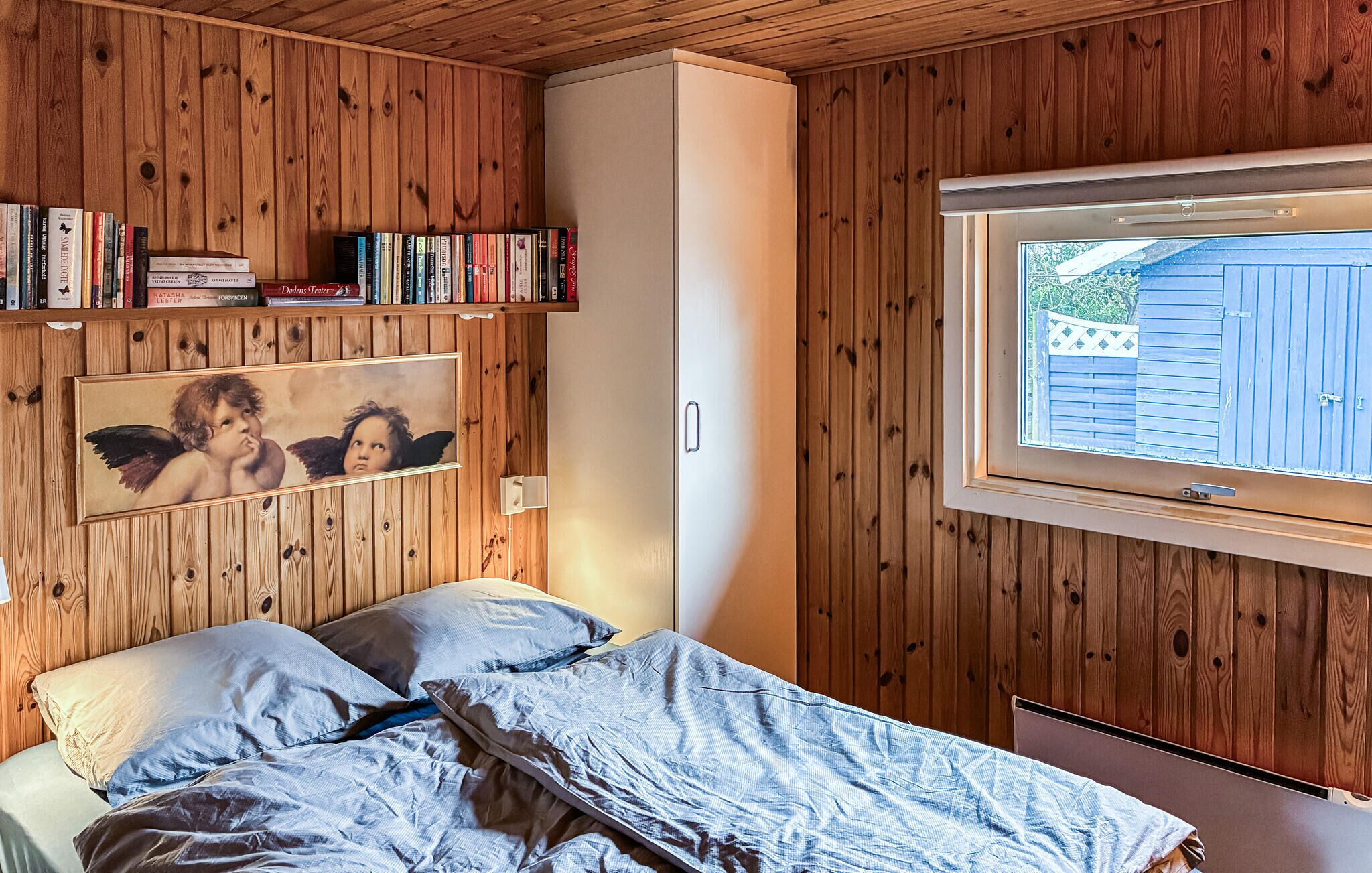 A bedroom with wooden walls, a bed with blue bedding, a bookshelf with books, a framed picture on the wall, and a window with a view of a wooden fence.