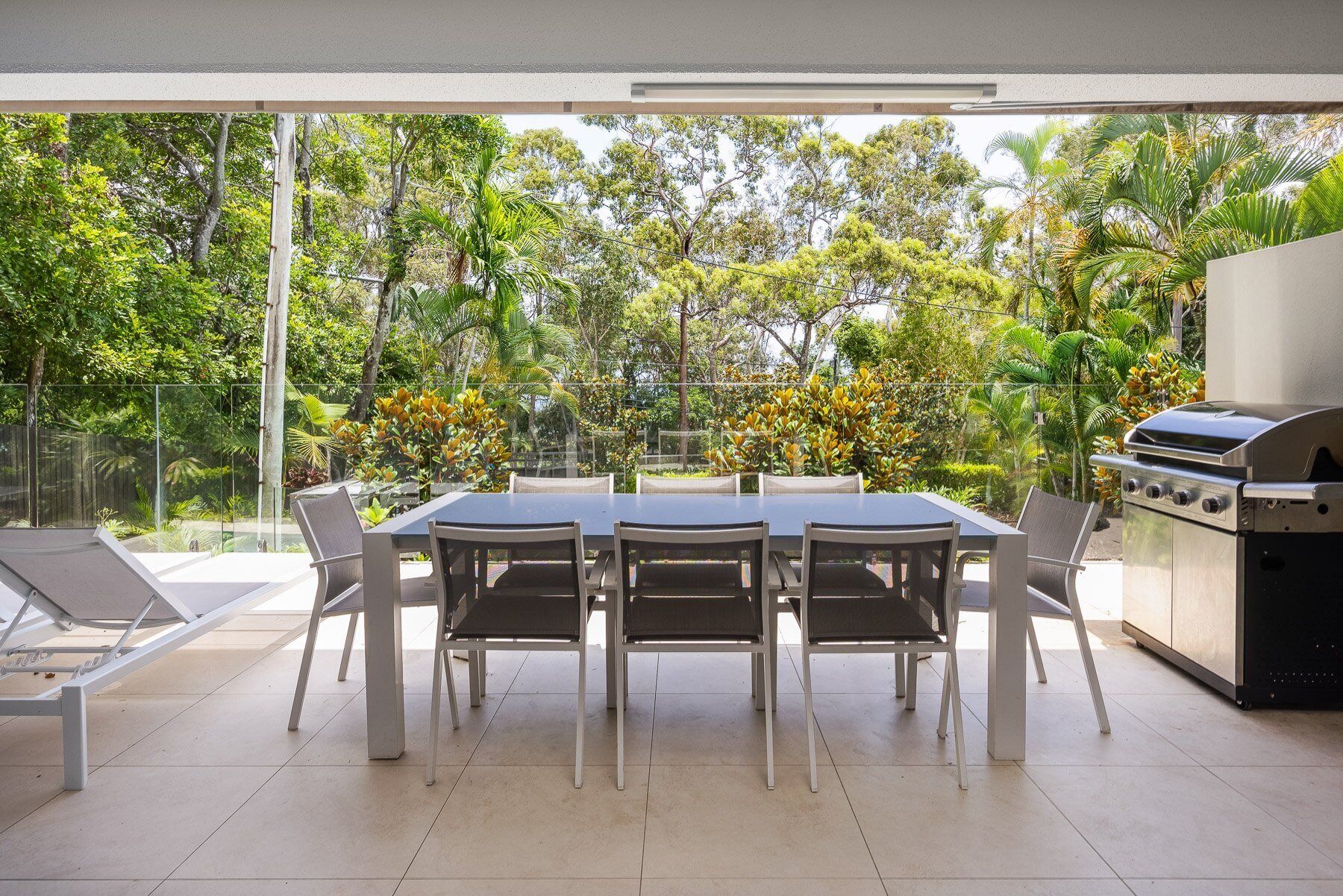 An outdoor patio with a dining table and chairs, a grill, and a view of a lush garden.