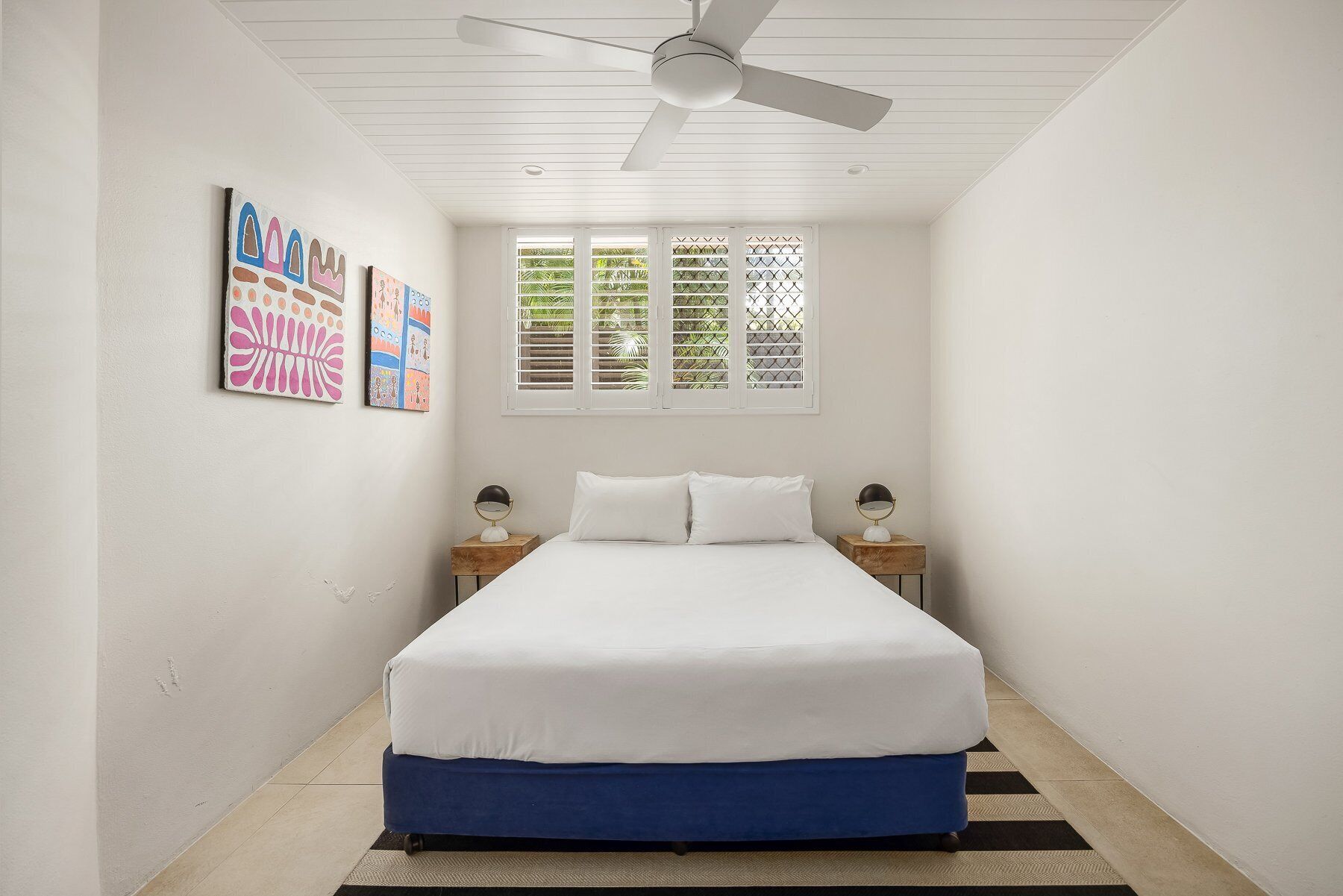 Une chambre à coucher avec un lit, deux tables de chevet, un ventilateur de plafond et une fenêtre avec des stores.